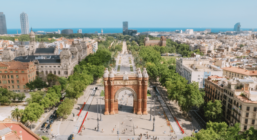 Aerial view of the Arc de Triomf in Barcelona, with tree-lined paths, surrounding city buildings, and the Mediterranean Sea in the background.