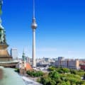 Cityscape of Berlin featuring the Fernsehturm TV tower, historical buildings, and greenery under a clear blue sky.