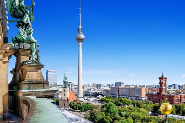 Cityscape of Berlin featuring the Fernsehturm TV tower, historical buildings, and greenery under a clear blue sky.