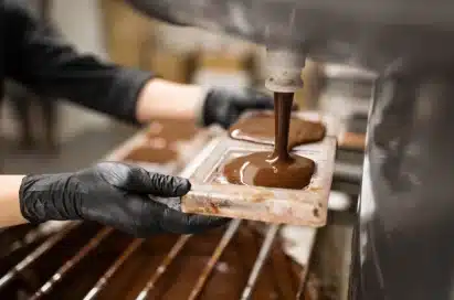 Person wearing black gloves pours melted chocolate into a mould, likely in a chocolate factory or workshop setting.