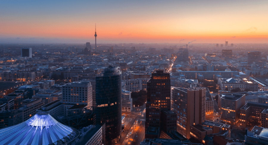 Aerial view of a city skyline at sunset with illuminated buildings, including a marquee-like structure in the foreground and a tall TV tower in the distance.