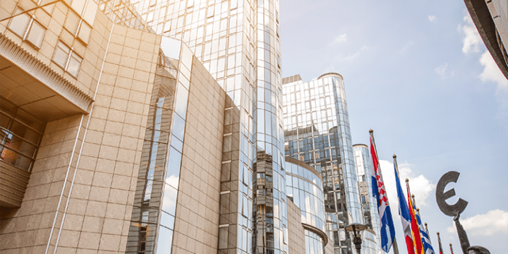 Modern glass and stone office buildings with several European national flags and a large euro currency sculpture in the foreground under a blue sky.