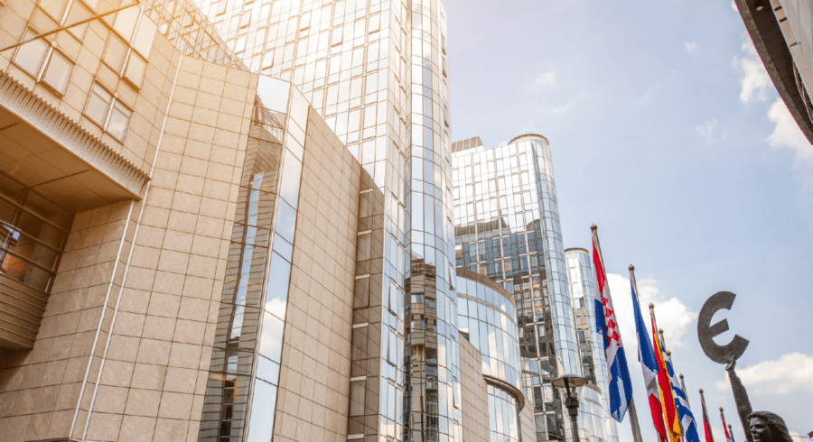 Modern glass and stone office buildings with several European country flags and a large euro symbol sculpture in front, under a partly cloudy sky.