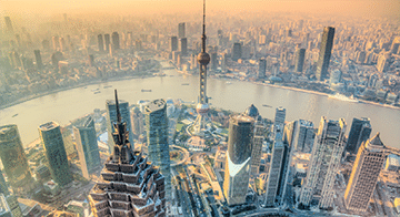 Aerial view of a modern city skyline with tall skyscrapers, a wide river, and a prominent observation tower in the centre, under a hazy sky.