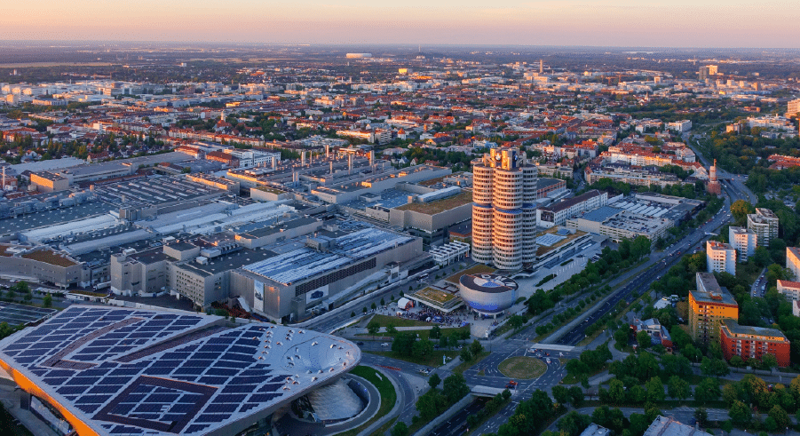 Aerial view of a large industrial complex with modern buildings and a tall round tower, surrounded by city streets and residential areas at sunset.
