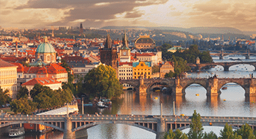 A cityscape of Prague shows multiple historic bridges crossing the Vltava River, with colourful buildings and church domes under a cloudy sky.