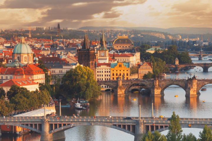 A panoramic view of Prague, showing the Vltava River, historic bridges, and colourful buildings under a partly cloudy sky at sunset.