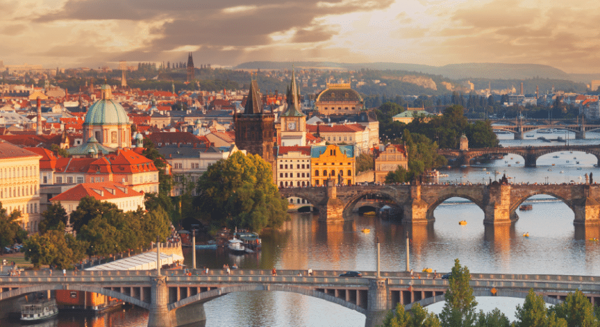 A panoramic view of Prague, showing the Vltava River, historic bridges, and colourful buildings under a partly cloudy sky at sunset.