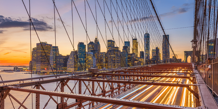 Brooklyn Bridge at sunset with light trails from traffic and the New York City skyline in the background.