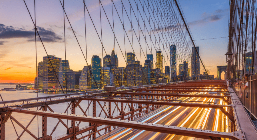 Brooklyn Bridge with light trails from traffic at sunset, Manhattan skyline in the background, and illuminated city buildings.