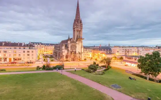 A large, ornate church with a tall spire stands in a city square surrounded by buildings and green lawns, illuminated by streetlamps at dusk.