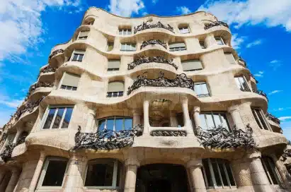 A multi-storey stone building with undulating façades and wrought iron balconies, characteristic of modernist architecture, shown against a blue sky with clouds.