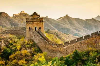 The Great Wall of China stretches across hilly terrain under a golden sky, with watchtowers visible along its length and greenery in the foreground.