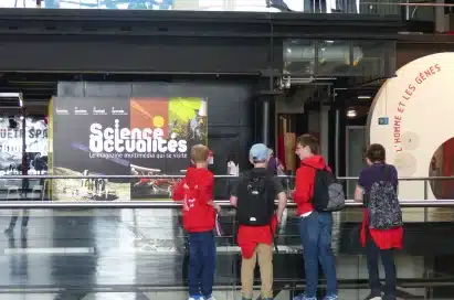 Four people with rucksacks stand indoors facing a large "Sciences Occultées" exhibit display at a science museum.