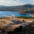 Aerial view of the Temple of Poseidon ruins on a hill overlooking the sea, with boats in the water and mountains in the background.
