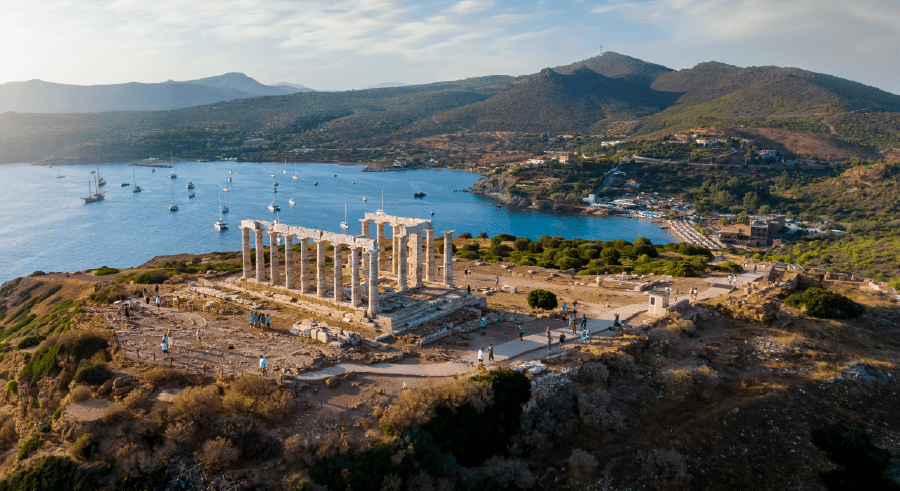 Aerial view of the ancient Temple of Poseidon ruins on a hill overlooking the sea, with boats anchored in the bay and surrounding hills in the background.