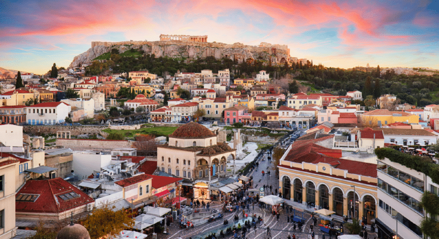 A view of the Acropolis on a hill above Athens, Greece, with colourful buildings and a busy city square in the foreground at sunset.