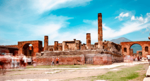 Ancient ruins of Pompeii with stone columns and structures under a bright blue sky; blurred figures of visitors are present in the foreground.
