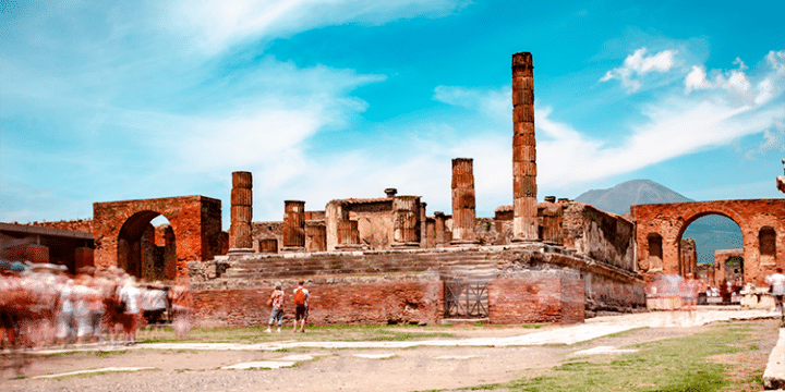 Ancient ruins of Pompeii with stone columns and structures under a bright blue sky; blurred figures of visitors are present in the foreground.