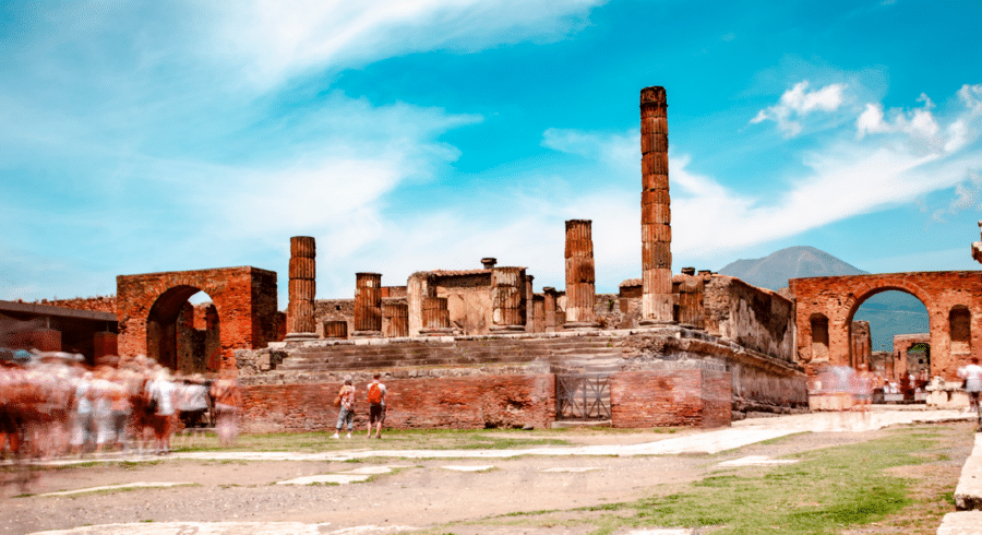 Ancient ruins of Pompeii with columns and brick structures under a blue sky, Mount Vesuvius in the background, and blurred crowds of visitors in the foreground.