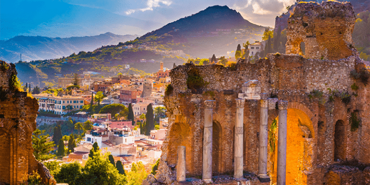 Ruins of an ancient stone amphitheatre in the foreground with a colourful hillside town and mountains in the background under a partly cloudy sky.