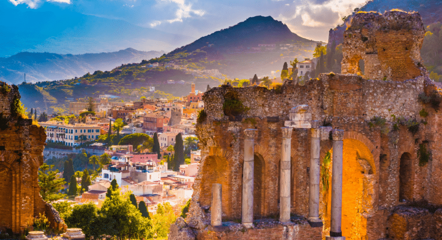 Ruins of an ancient stone theatre with columns in the foreground, colourful town buildings and green hills in the background under a partly cloudy sky.
