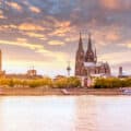 View of Cologne’s skyline at sunset, featuring the Cologne Cathedral, nearby churches, and the Rhine River in the foreground with boats on the water.