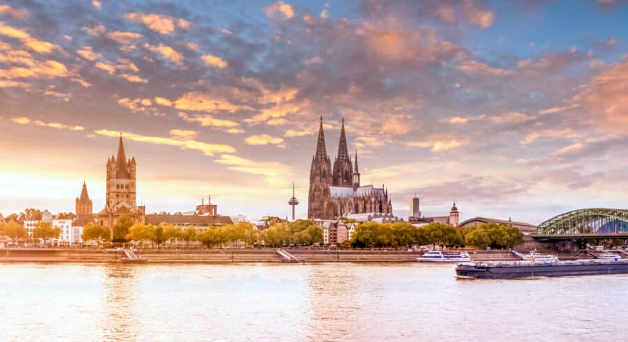 Skyline of Cologne, Germany at sunset, featuring the Cologne Cathedral, the Great St Martin Church, and the River Rhine with boats.