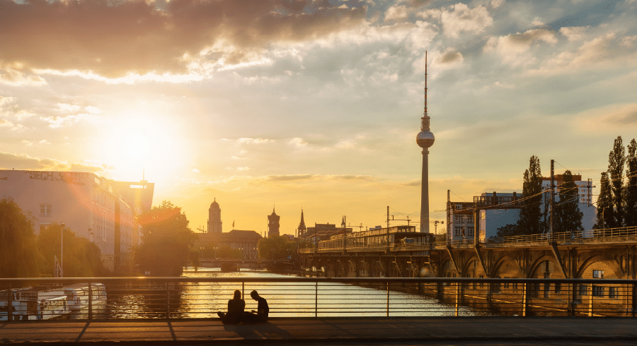 A sunset view of Berlin with the TV tower, railway tracks, and Spree River; two people sit by the riverside in the foreground.