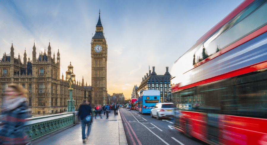 People and vehicles cross Westminster Bridge in London with Big Ben and the Houses of Parliament in the background; a red double-decker bus is blurred in motion.