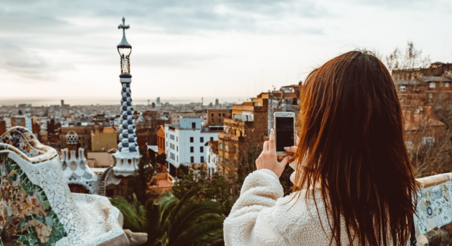 A person in a light coat takes a photo of the cityscape and tiled structures at Park Güell in Barcelona, Spain, during an overcast day.