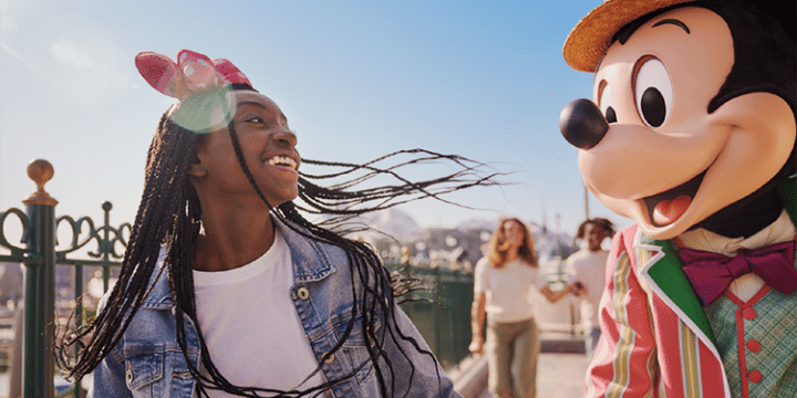 A girl smiling and walking outdoors beside a person in a Mickey Mouse costume wearing a colourful suit and hat, with other people in the background.