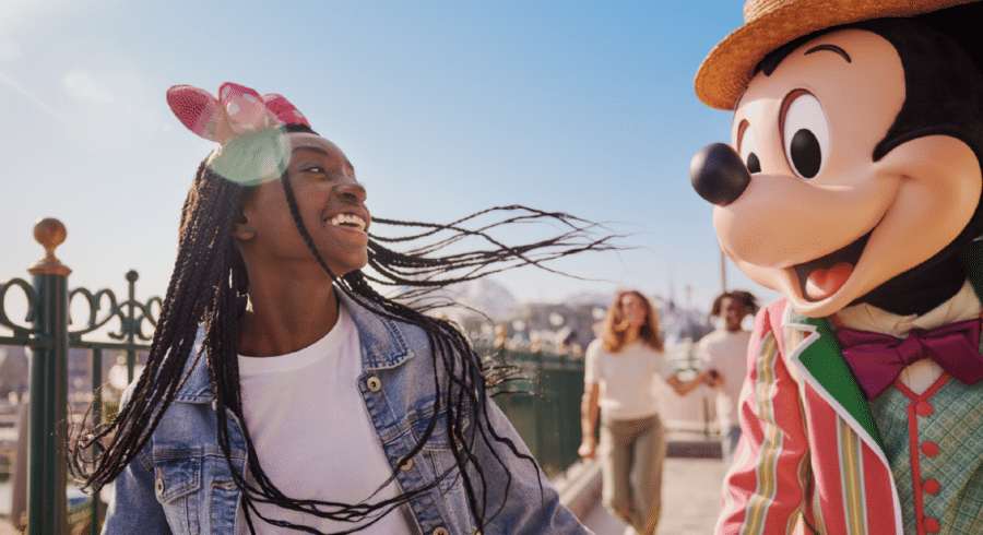 A girl in a denim jacket smiles and looks at a person in a Mickey Mouse costume at an outdoor amusement park.