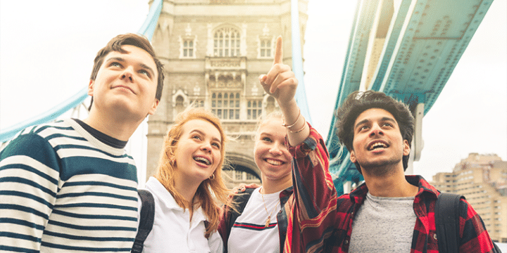 Four young people stand together smiling, with one pointing upwards, in front of Tower Bridge in London on a sunny day.
