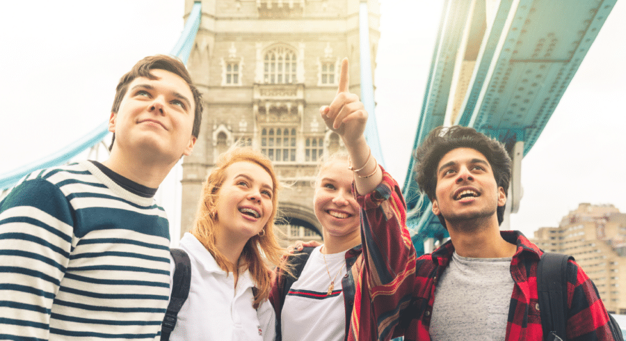 Four young people stand together, smiling and looking up, with one person pointing. They are outdoors near a large stone bridge structure.