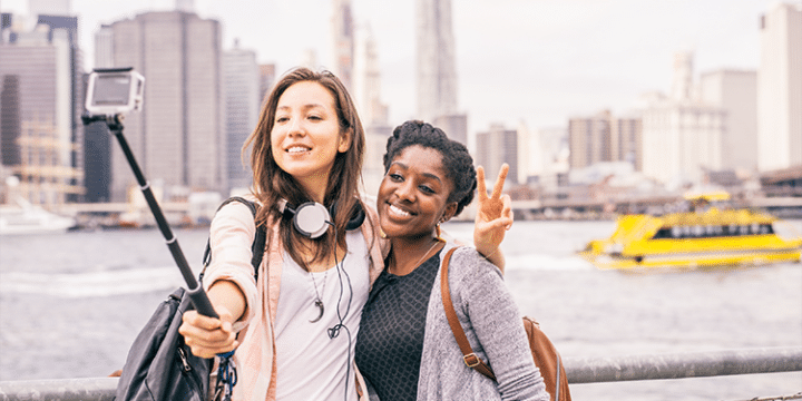 Two young women take a selfie with a selfie stick by a waterfront, with a city skyline and a yellow ferry in the background.