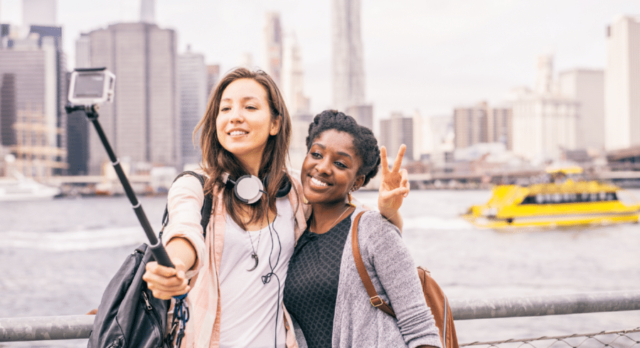 Two women standing by a waterfront take a selfie with a mobile on a stick; city skyline and a yellow ferry are visible in the background.