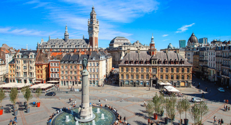 A city square with a fountain, historic buildings, and a tall clock tower under a clear blue sky. People are walking and sitting around the square.