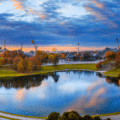 A panoramic view of a park with a lake, surrounded by autumn trees and unique tent-like stadium structures under a partly cloudy sky at sunset.
