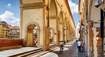 People walk and cycle along a narrow street lined with historic arches and buildings under a bright, partly cloudy sky.