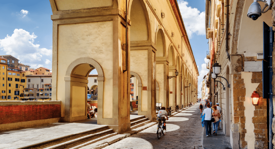 People walk and ride bicycles along the covered Vasari Corridor in Florence, Italy, with arches casting shadows on the pavement under a bright, partly cloudy sky.