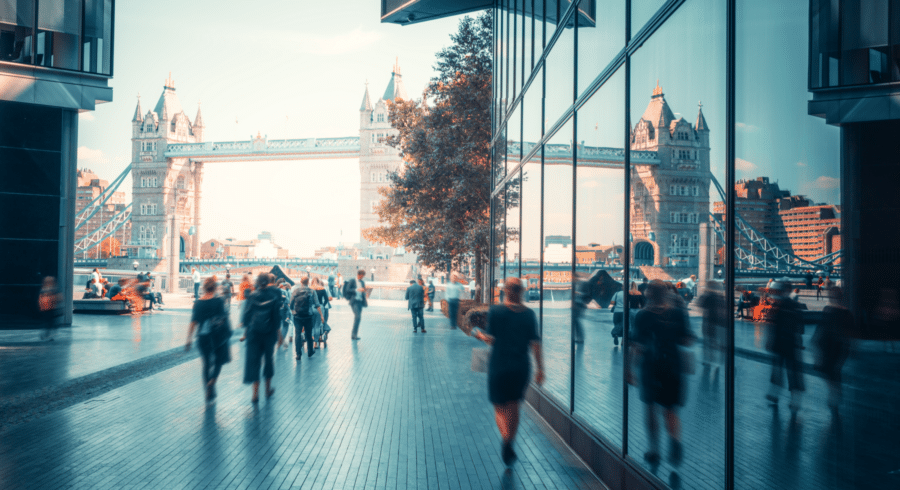 People walk along a modern riverside path with reflections on glass panels; Tower Bridge is visible in the background under a clear sky.