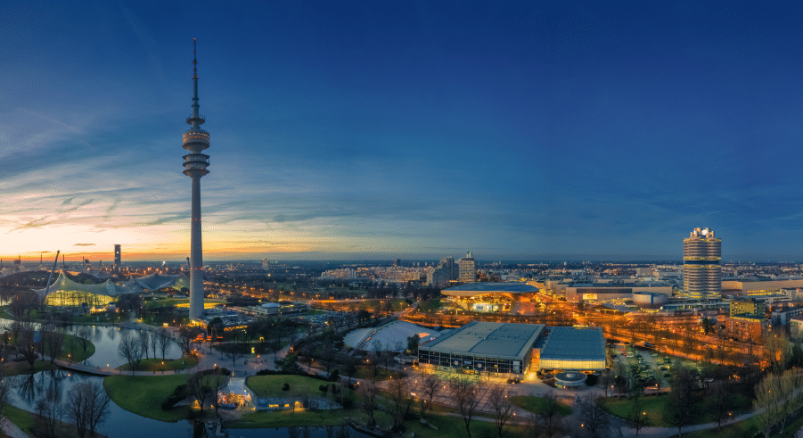 Panoramic view of Munich's Olympiapark at dusk, featuring the Olympic Tower, illuminated buildings, and park areas with a sunset sky in the background.