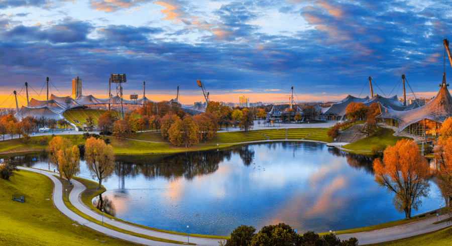 A panoramic view of a park with a large pond, pathways, and distinctive tent-like structures under a partly cloudy sky at sunset.