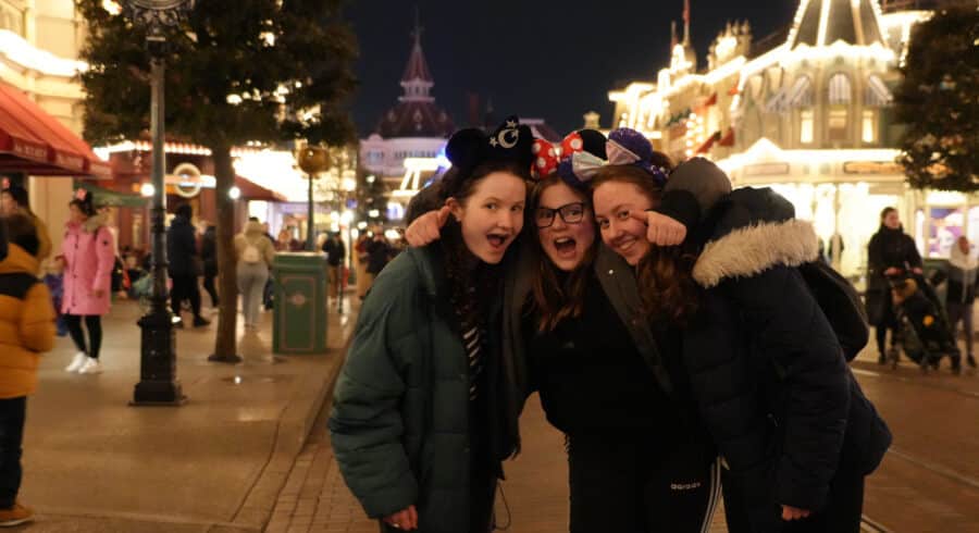Three girls wearing mouse ears pose smiling and hugging on a brightly lit street at night in a theme park with people walking in the background.