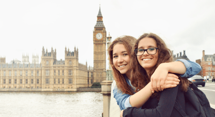 Two young people smiling and hugging on a bridge with the Houses of Parliament and Big Ben in London visible in the background.