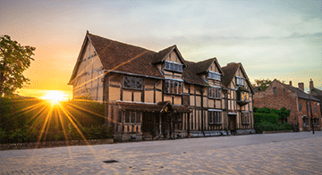 A historic timber-framed house with a steep pitched roof at sunset, with the sun shining through trees on the left and a cobbled street in front.
