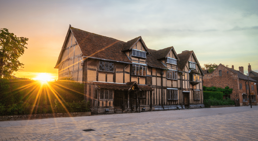 An historic, timber-framed house with leaded windows, photographed at sunset with the sun visible on the left and a clear sky above.