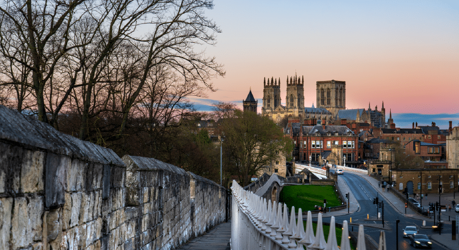 View of York city wall leading towards York Minster at sunset, with historic buildings and trees visible along the street.