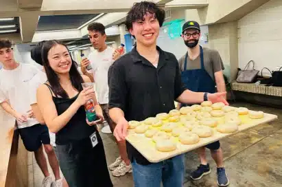 A group of young adults stands indoors; one person at the front holds a tray of bagels and smiles at the camera, while others stand nearby, some holding drinks.